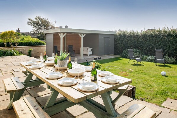 Un jardin avec une table de pique-nique en bois, prête pour un repas, un espace de détente abrité et une pelouse bien entretenue.