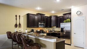A modern kitchen with dark cabinets, stainless steel appliances, and a bar area with leather stools. - 1424ww - The Retreat at Championsgate (Davenport)
