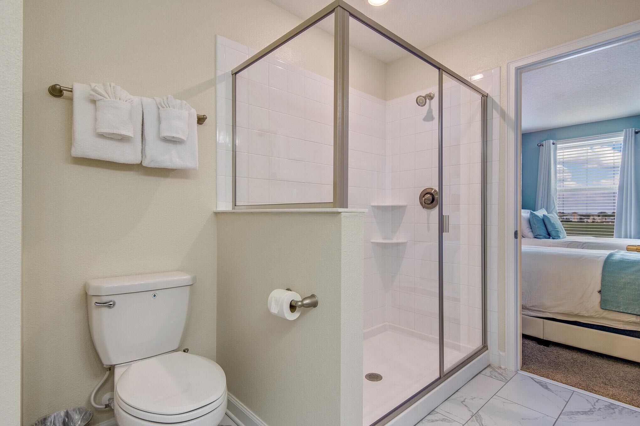 A bathroom with a toilet, a glass-enclosed shower, and a visible bed in the adjacent room.