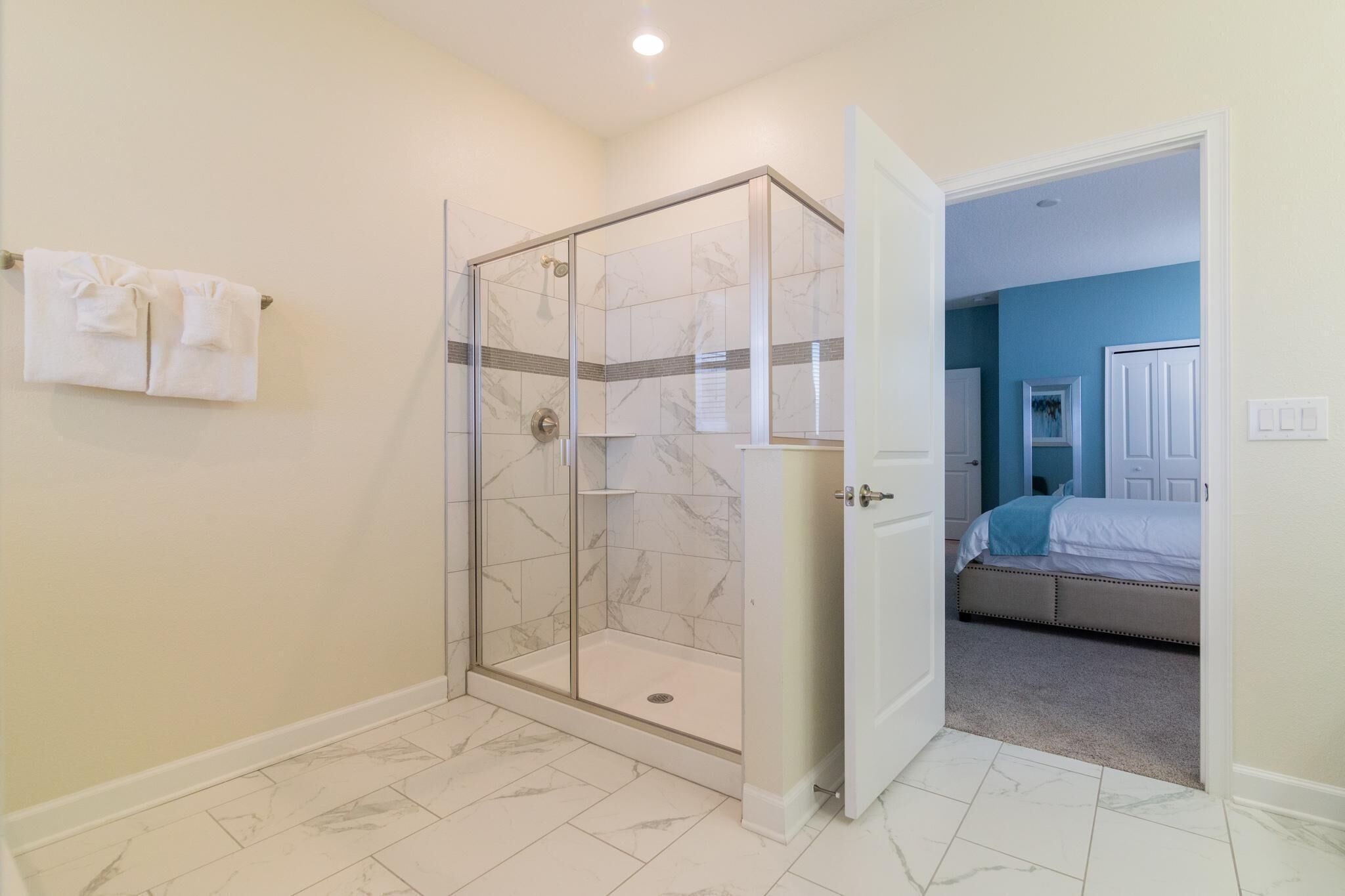 A bathroom with a glass-enclosed shower, a white door, and a visible bedroom with a bed and a blue wall.