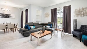 A modern living room with a sectional sofa, a coffee table, and a dining area with a table and chairs. - 8919sid-the Retreat at Championsgate (Davenport)