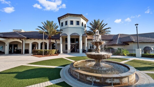A courtyard with a central fountain, surrounded by a paved walkway and green lawns, with a building featuring a tower and arched entryways in the background. - 1559pw NON Renting 8/15/21the Retreat at Championsgate (Davenport)