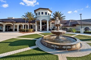 A courtyard with a central fountain, surrounded by a paved walkway and green lawns, with a building featuring a tower and arched entryways in the background. - 1559pw NON Renting 8/15/21the Retreat at Championsgate (Davenport)