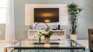 A modern living room with a glass coffee table, a flat-screen TV mounted on a textured wall, a white console table with decorative items, and a tall potted plant. - 1559pw NON Renting 8/15/21the Retreat at Championsgate (Davenport)