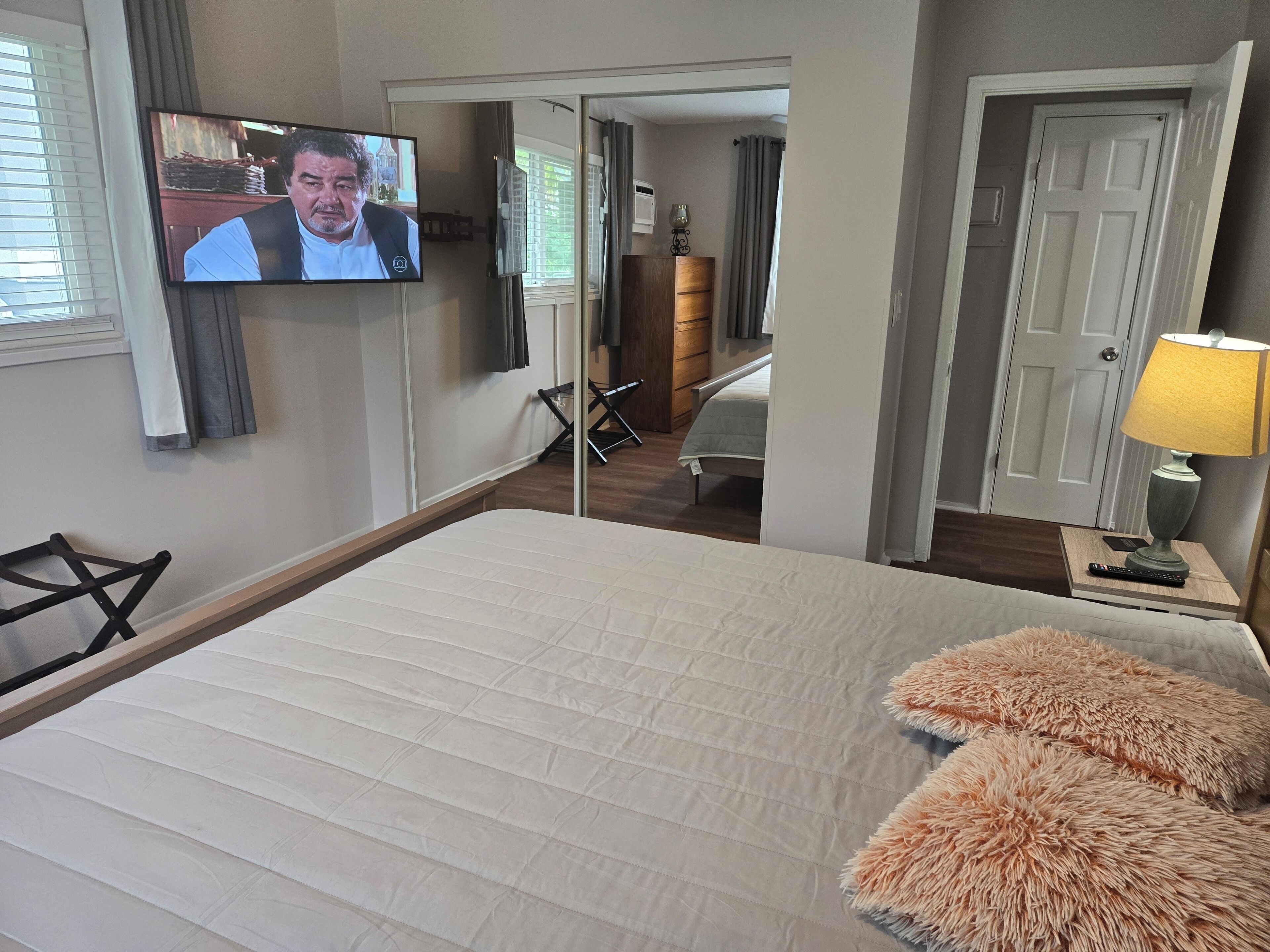 Habitación de hotel con cama, televisor montado en la pared, lámpara en una mesita de noche y una puerta que da a otra habitación.