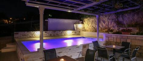 Un área de piscina al aire libre con una pérgola, asientos y una pared de piedra.
