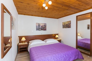 A bedroom with a wooden ceiling, a purple bedspread, two bedside lamps, and two paintings on the wall.