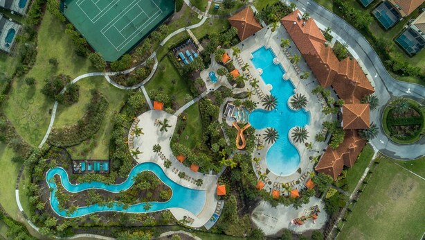 Eine Vogelperspektive zeigt ein Resort mit mehreren Schwimmbecken, einem Tennisplatz und begrünten Anlagen.
