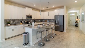 A modern kitchen with white cabinets, granite countertops, a stainless steel refrigerator, and a central island with bar stools. - 1559fd - The Retreat at Championsgate (Davenport)