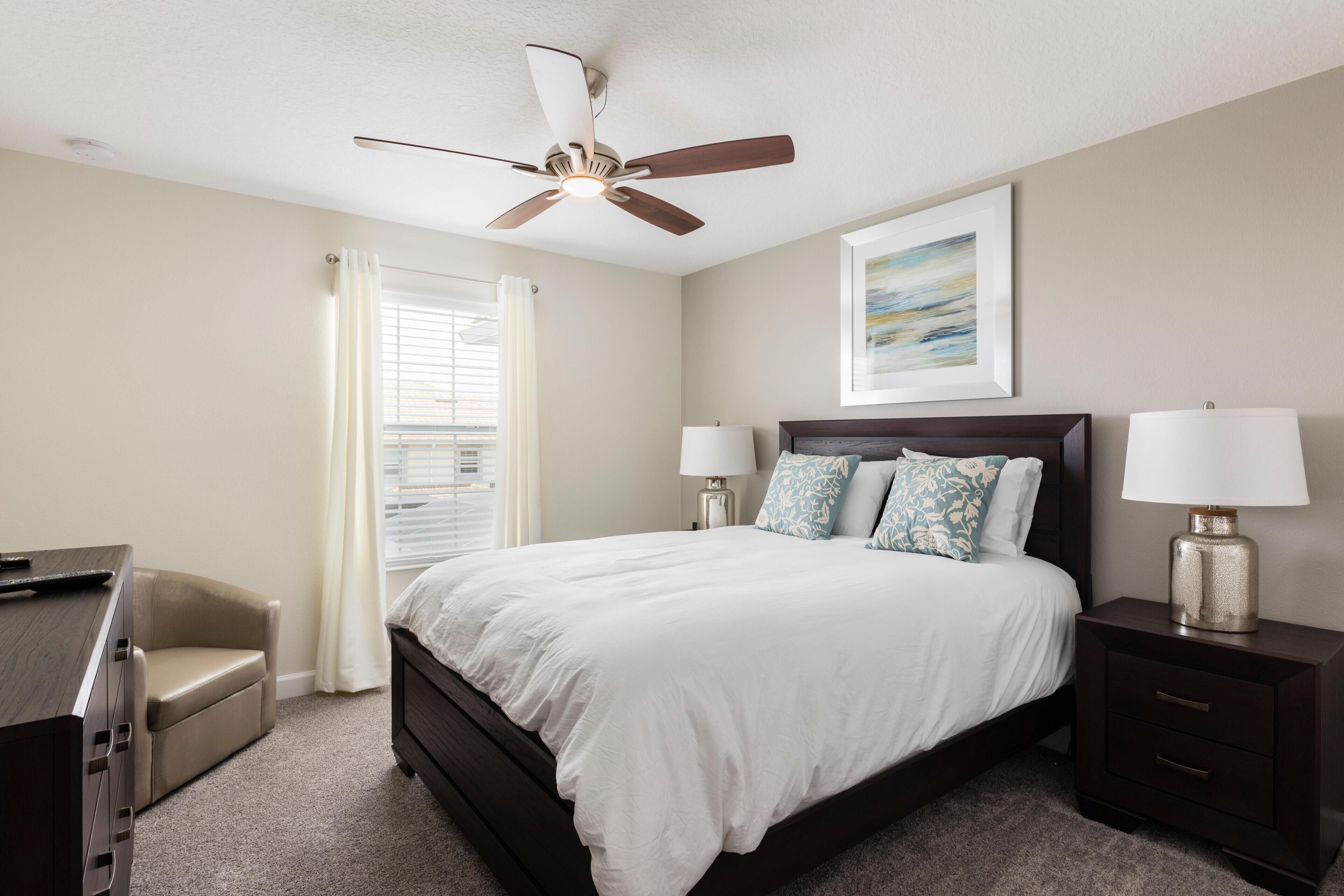 A bedroom with a bed, two bedside tables with lamps, a ceiling fan, a window with curtains, and a desk.