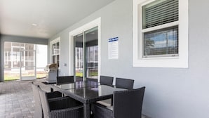 Un patio techado con una mesa de vidrio y sillas de mimbre, un letrero que indica las normas de la piscina, y vista de la piscina a través de puertas de cristal.