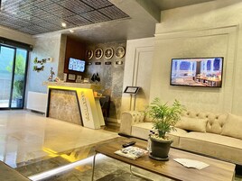 Zona de recepción de hotel con mostrador, sofá, mesa baja con una revista, planta en maceta y televisor fijado en la pared.