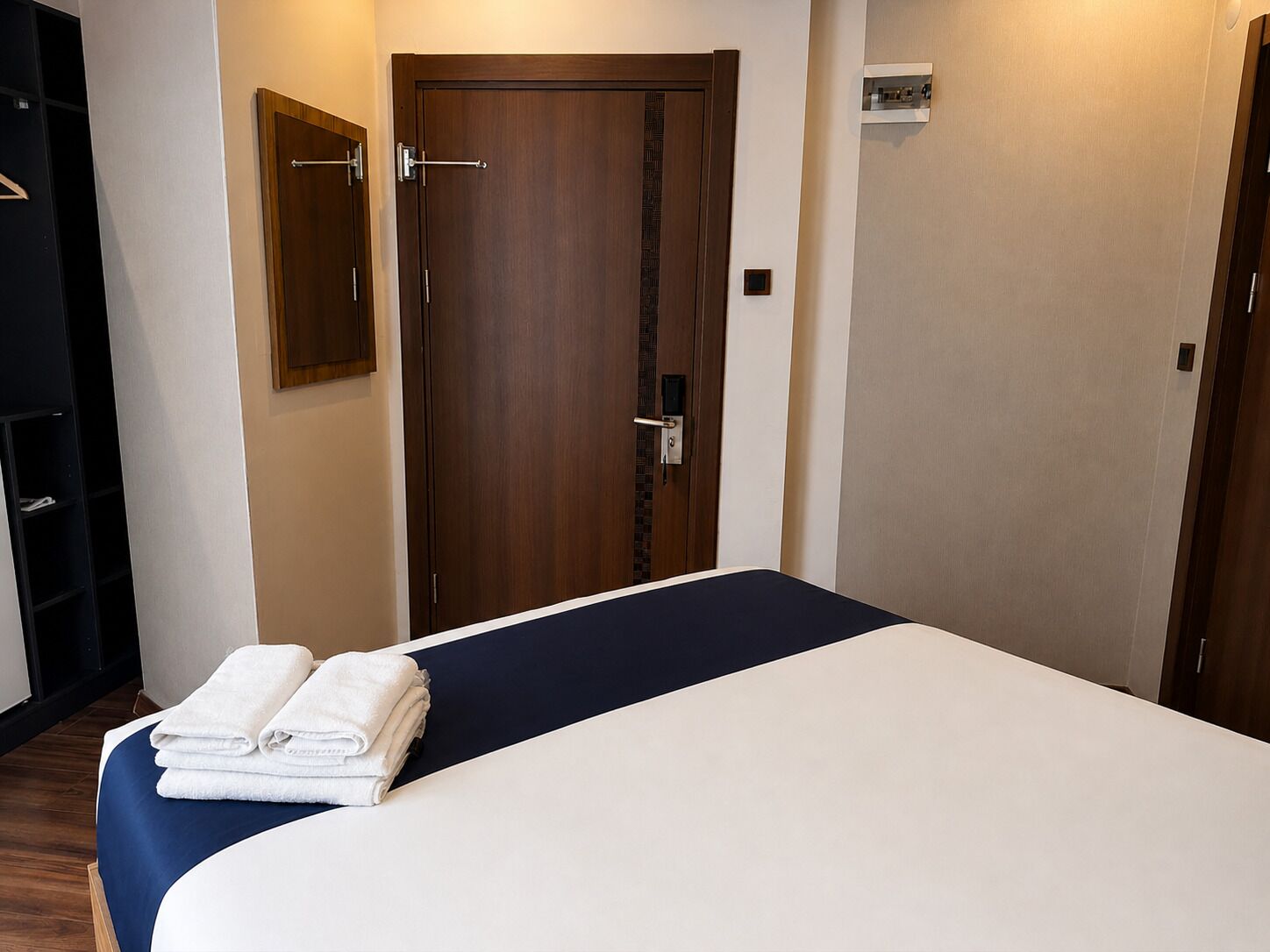 A hotel room with a bed, a wooden door, a small shelf, and a wall-mounted light.