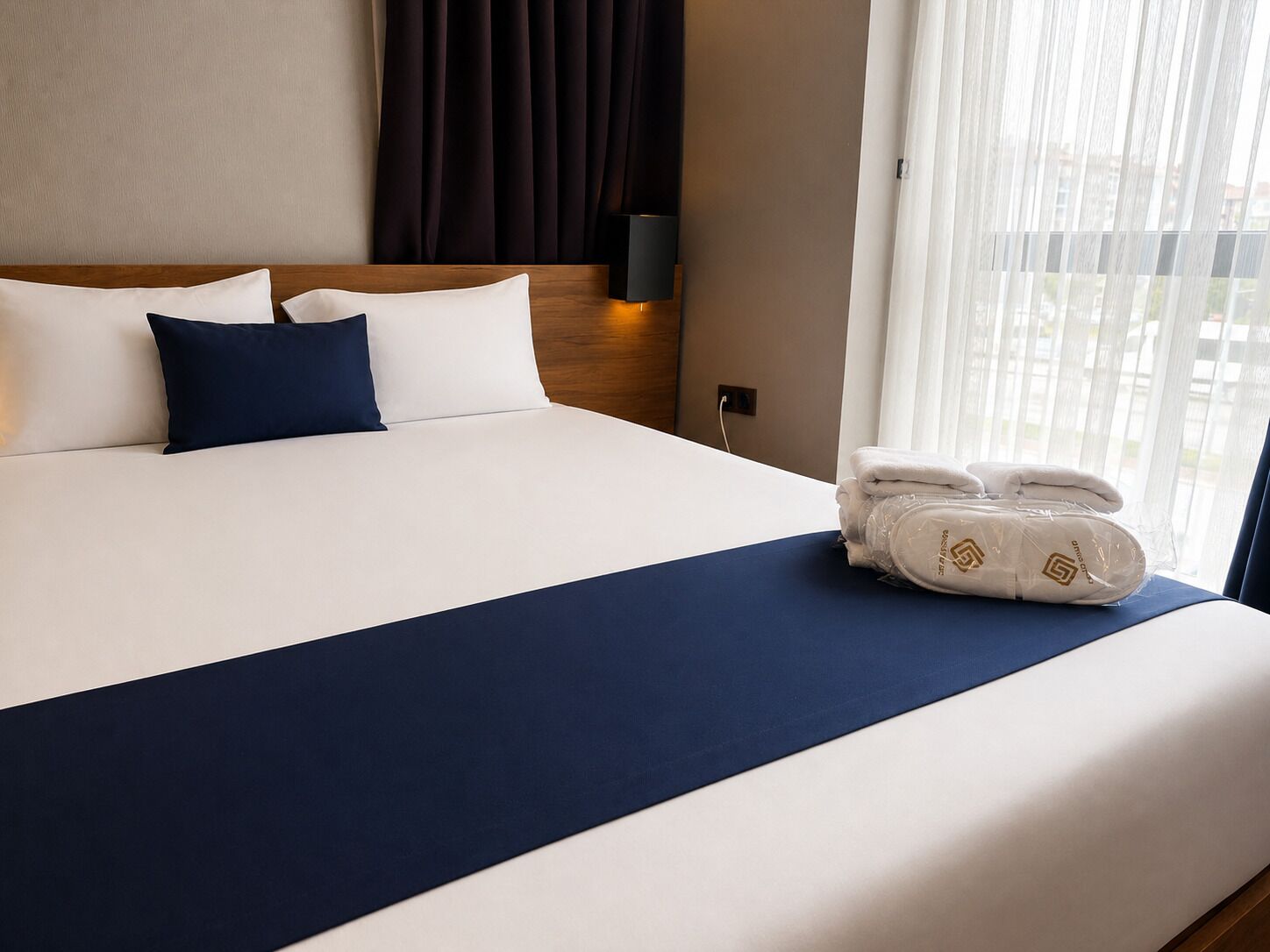 Una cama bien hecha con ropa de cama blanca y azul marino, un cabecero oscuro y una ventana con cortinas translúcidas.