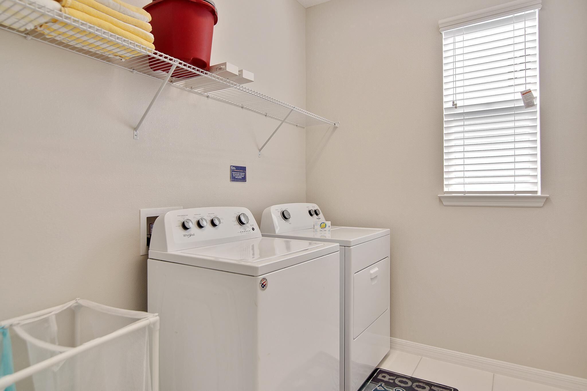 A laundry room with two white front-loading washers and dryers, a metal drying rack, a red water heater, and a window with blinds.
