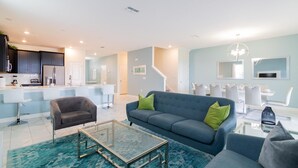 Een moderne woonkamer met een bank, fauteuil en salontafel. In de achtergrond is een keuken zichtbaar met donkere kasten en witte apparaten.