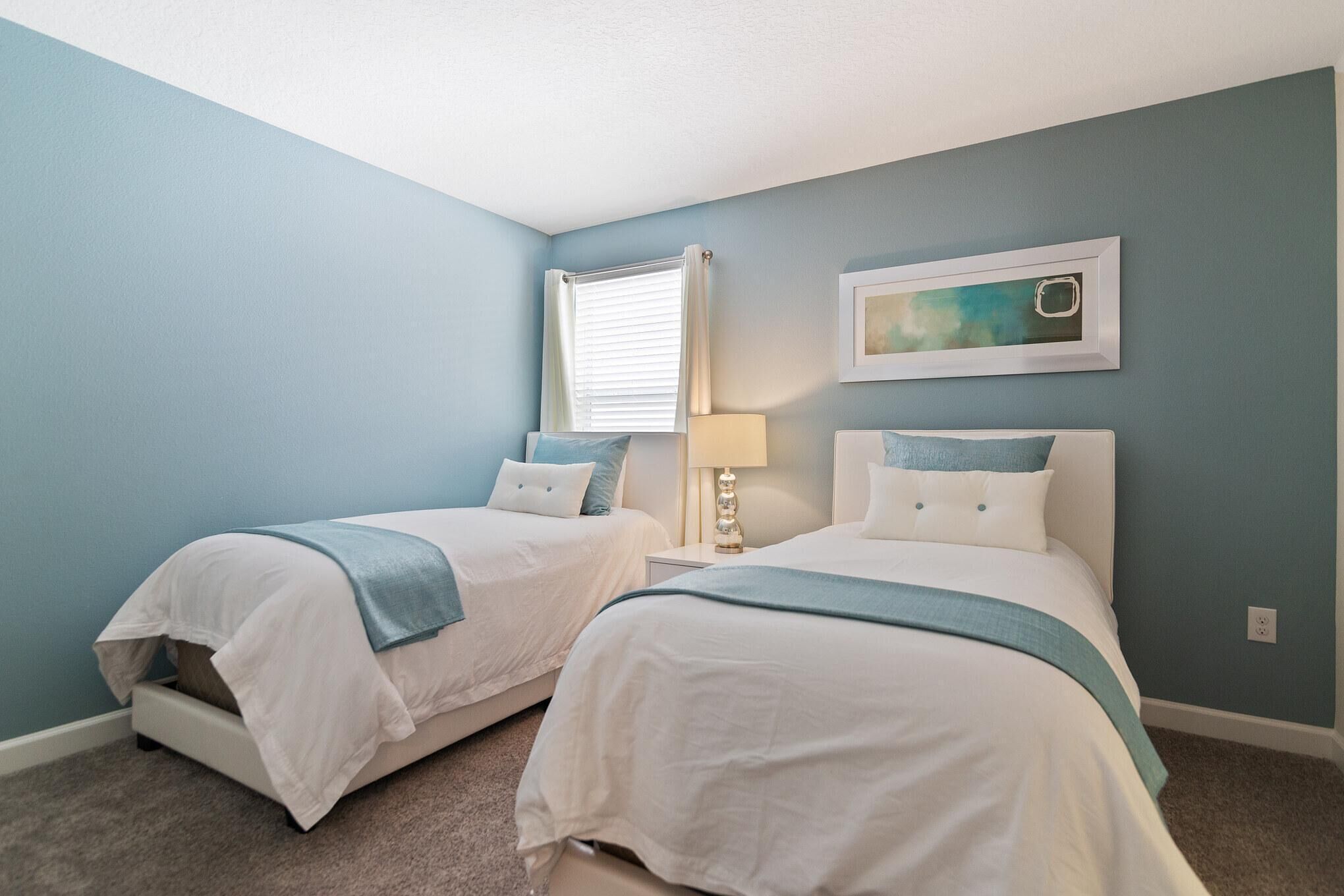 A bedroom with two beds, a window with curtains, a bedside lamp, and a framed picture on the wall.