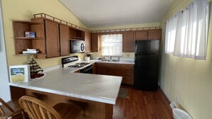 Une cuisine avec des meubles en bois, un réfrigérateur noir, une cuisinière et une fenêtre ornée de rideaux légers.