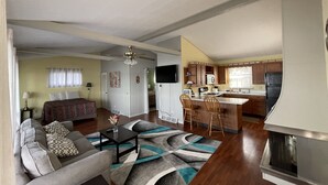 Un salon comprenant un canapé, une table basse et un tapis à motifs. On y trouve également un coin cuisine avec des meubles en bois et un réfrigérateur. Un ventilateur de plafond est visible dans le salon.