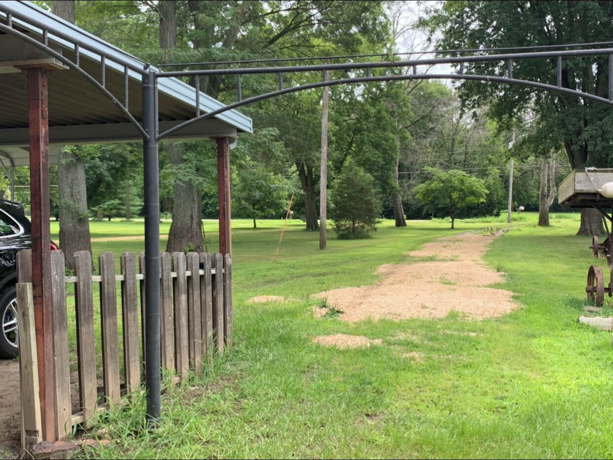 A covered outdoor area with a wooden fence, a dirt path, and trees in the background.