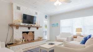 A living room with a white brick fireplace, a flat-screen TV mounted above it, and a comfortable seating area with white sofas and a coffee table.