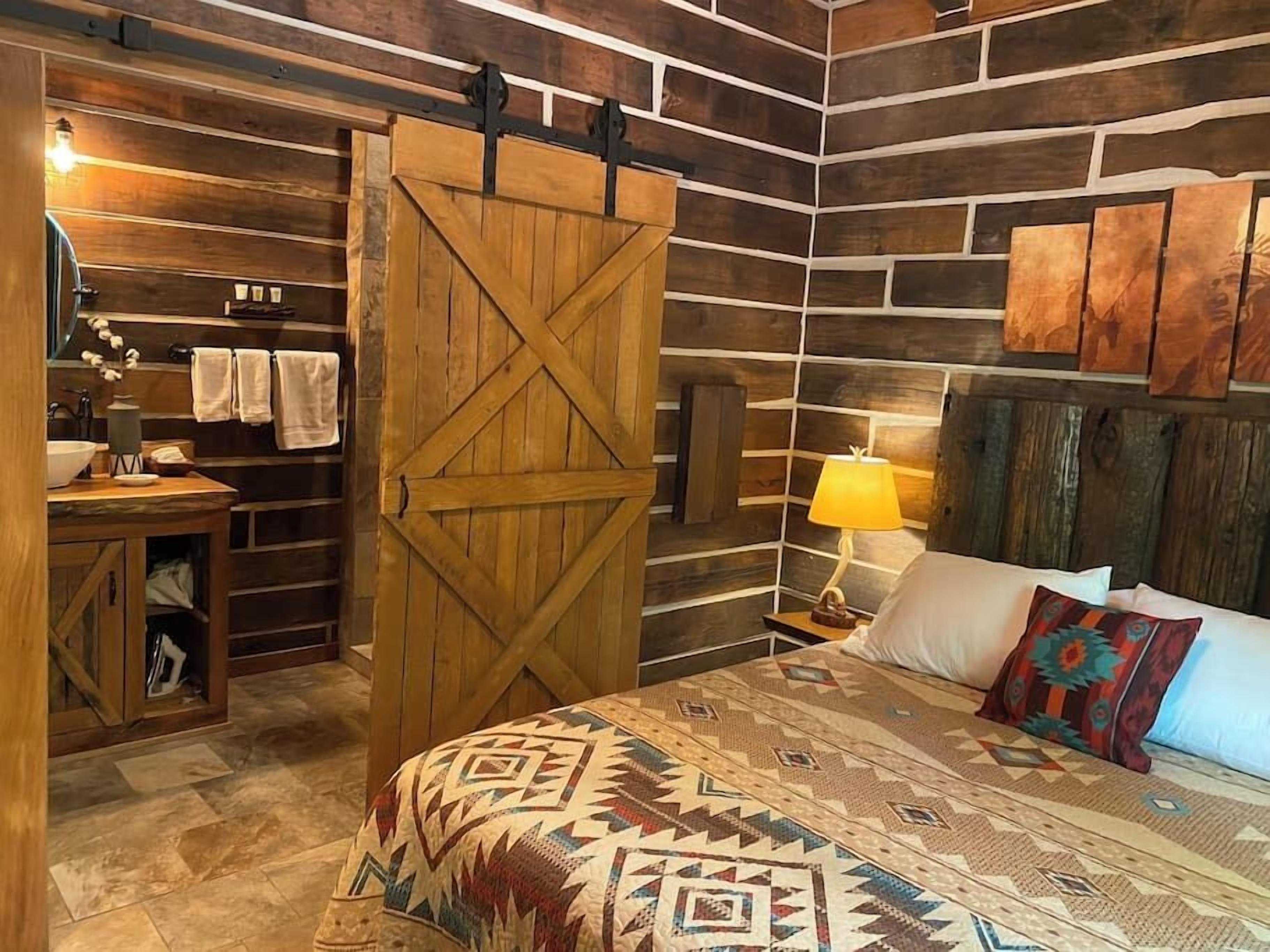 A rustic room with a wooden door, a bed with patterned bedding, a nightstand with a lamp, and a wooden wall with a shelf.