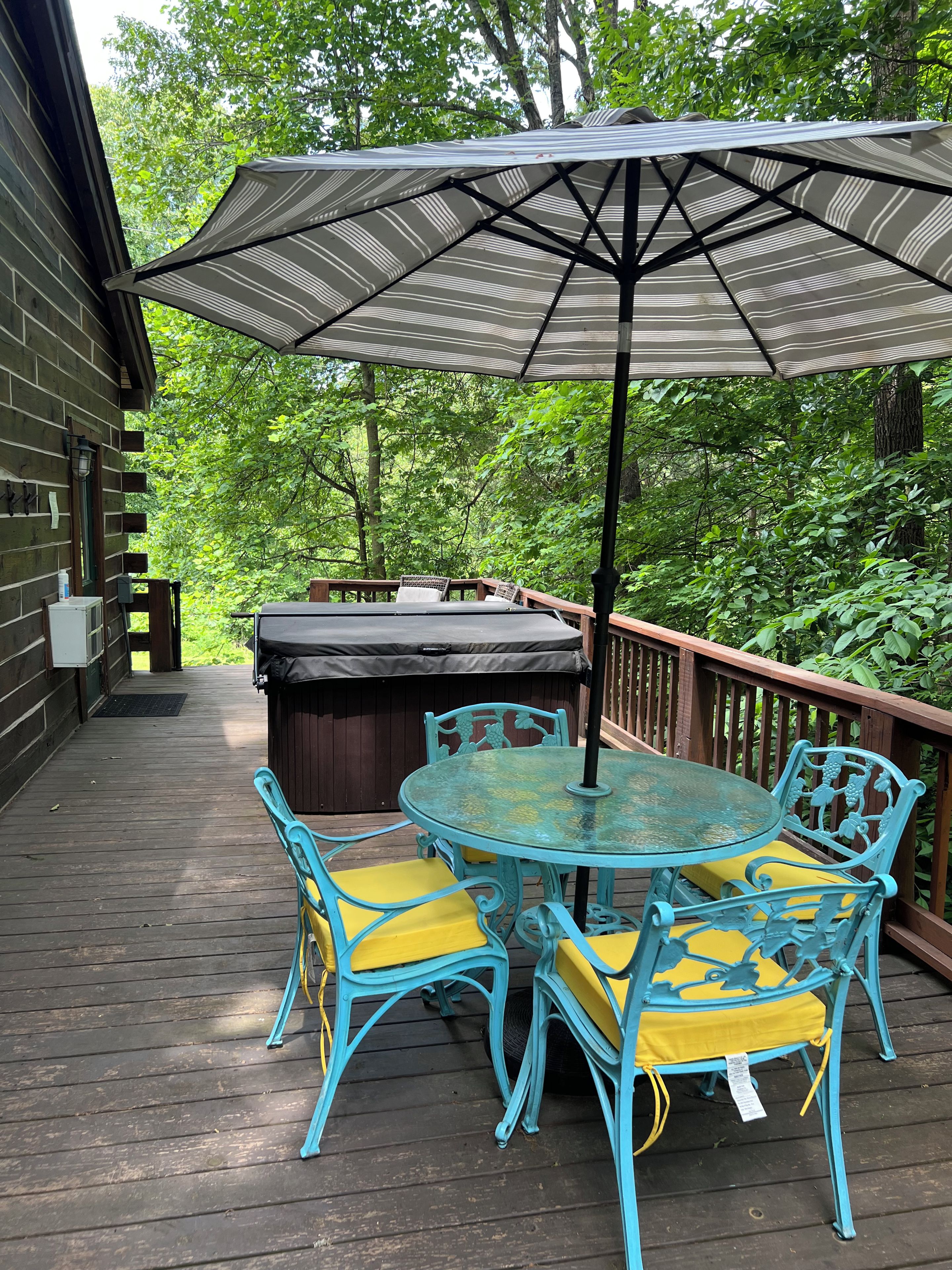 A wooden deck with a glass table and chairs, a large striped umbrella, and a hot tub.
