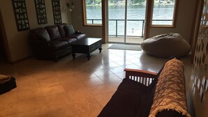Un salon avec un canapé marron foncé, une table basse en bois et un fauteuil en forme de poire. Une grande fenêtre offre une vue sur un lac et des maisons.