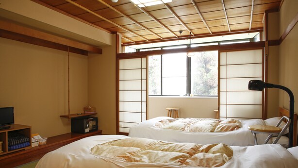 Una habitación tradicional japonesa con suelo de tatami, dos camas con ropa de cama blanca, un televisor sobre una estantería de madera y un ventanal con paneles shoji.