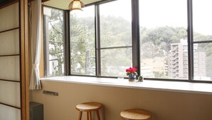 Una habitación con un amplio ventanal, dos taburetes de madera, una mesita con un florero y una lámpara colgante.