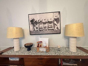 Ein Zimmer mit einem Holztisch, zwei Lampen, einem gerahmten Bild und einem Korb voller Gegenstände auf einem gemusterten Teppich.