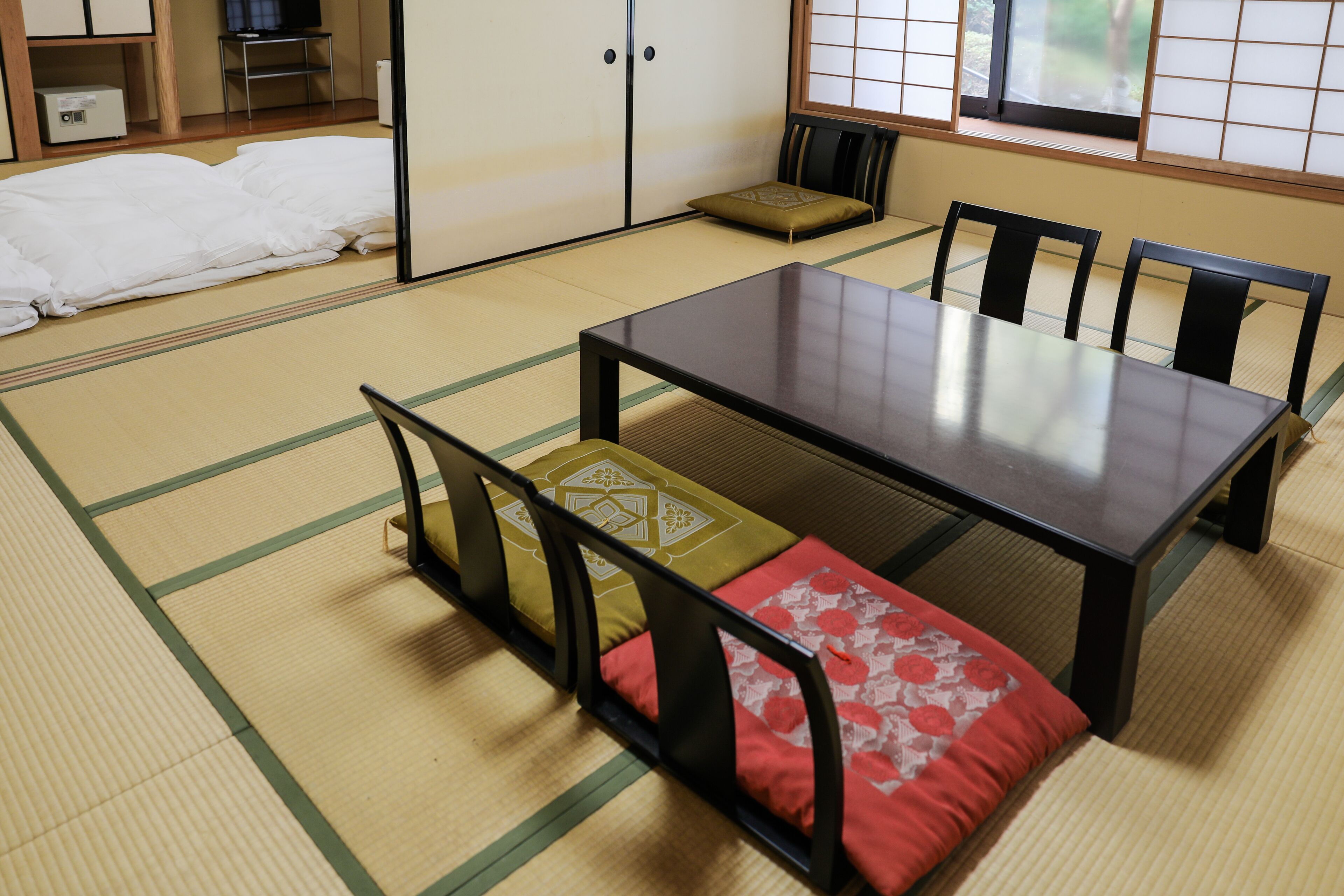 Una habitación con suelo de tatami, una mesa baja y sillas, una cama futón y puertas correderas.