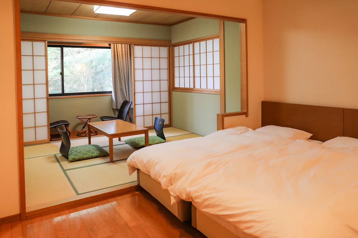 A traditional Japanese room with tatami flooring, a low wooden bed, a futon, a small table with a chair, and a sliding shoji screen.