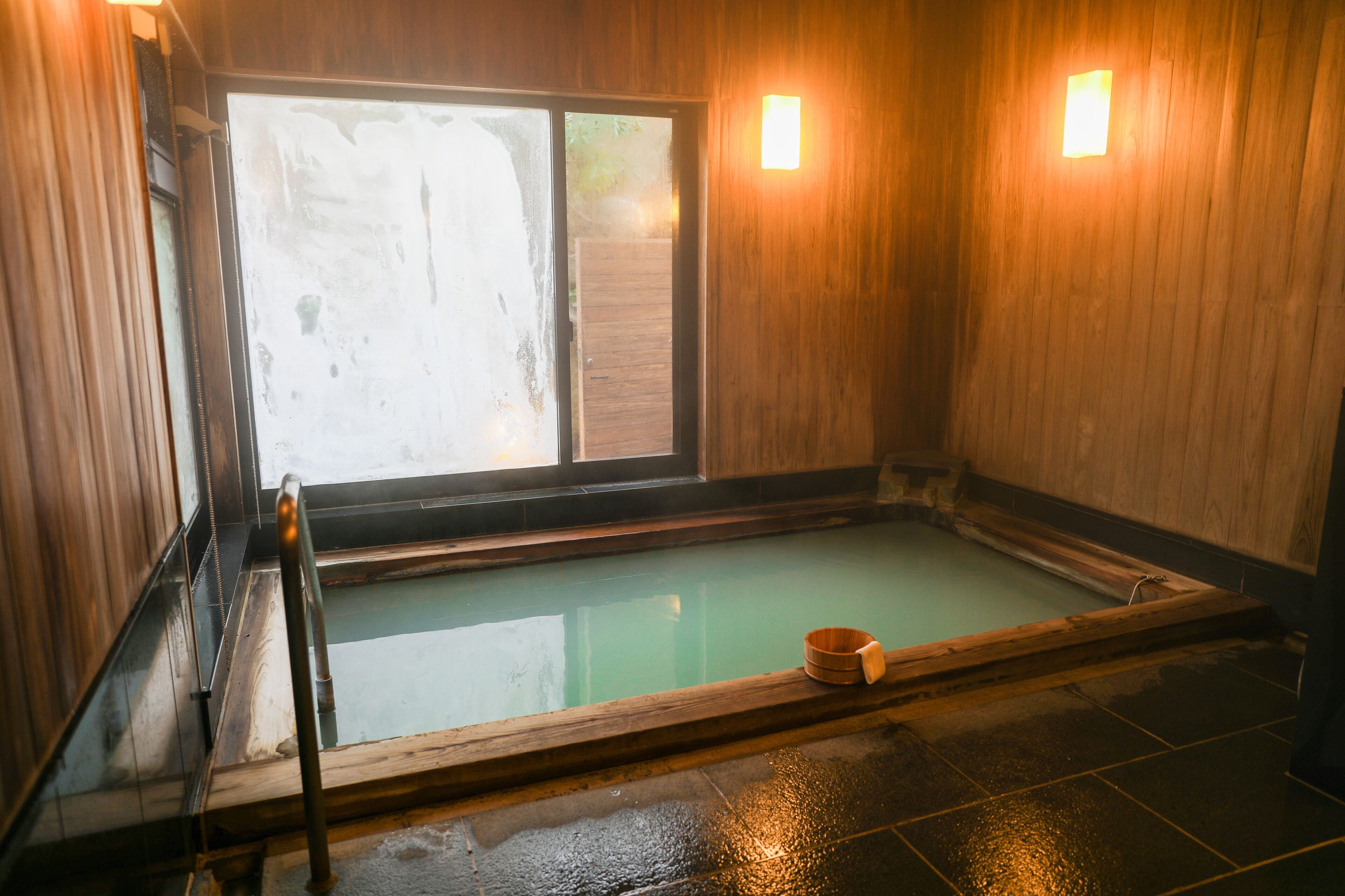 A traditional Japanese onsen with a wooden interior, a wooden tub, and a view of the outside.