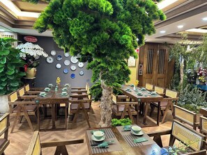 Restaurante com mesas e cadeiras de madeira, uma árvore central e uma parede decorada com pratos.