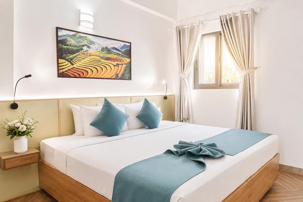 Une chambre à coucher bien rangée, avec un lit, des oreillers, une table de chevet sur laquelle se trouve un vase, un tableau accroché au mur et une fenêtre avec des rideaux.