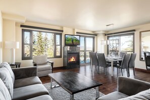 A modern living room with a fireplace, a dining area, and large windows offering a view of trees. - Ski-In/Out in Park City Village|Heated Pool|Hot Tub|Managed by Park City Lodging (Park City)