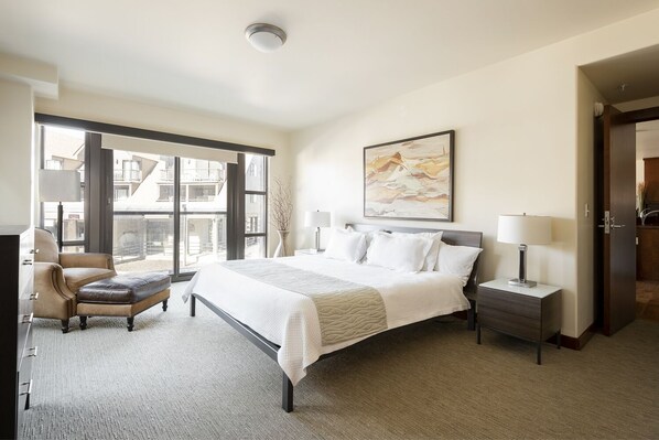 Une chambre d’hôtel moderne avec un grand lit, un coin salon avec un fauteuil, une table de chevet et un tableau au mur.