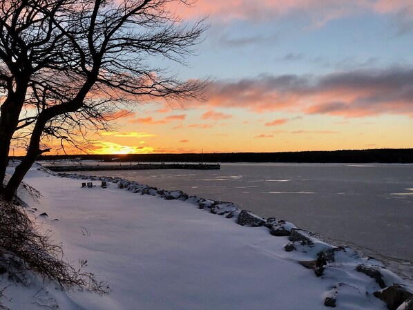 A frozen lake with a sunset in the background, surrounded by snow-covered ground and trees.