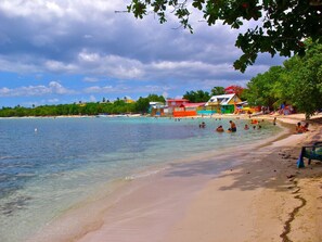 A beach with clear turquoise water, a sandy shore, and colorful buildings in the background.