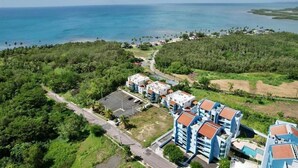 An aerial view of a coastal area with a cluster of buildings, a road, and a clear view of the ocean. - Spectacular Ocean View  Penthouse, Terrace, 2br, 2bth -  Surrounded by Nature (Cabo Rojo)