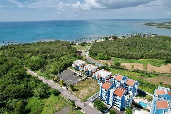 An aerial view of a coastal area with a cluster of buildings, a road, and a clear view of the ocean.