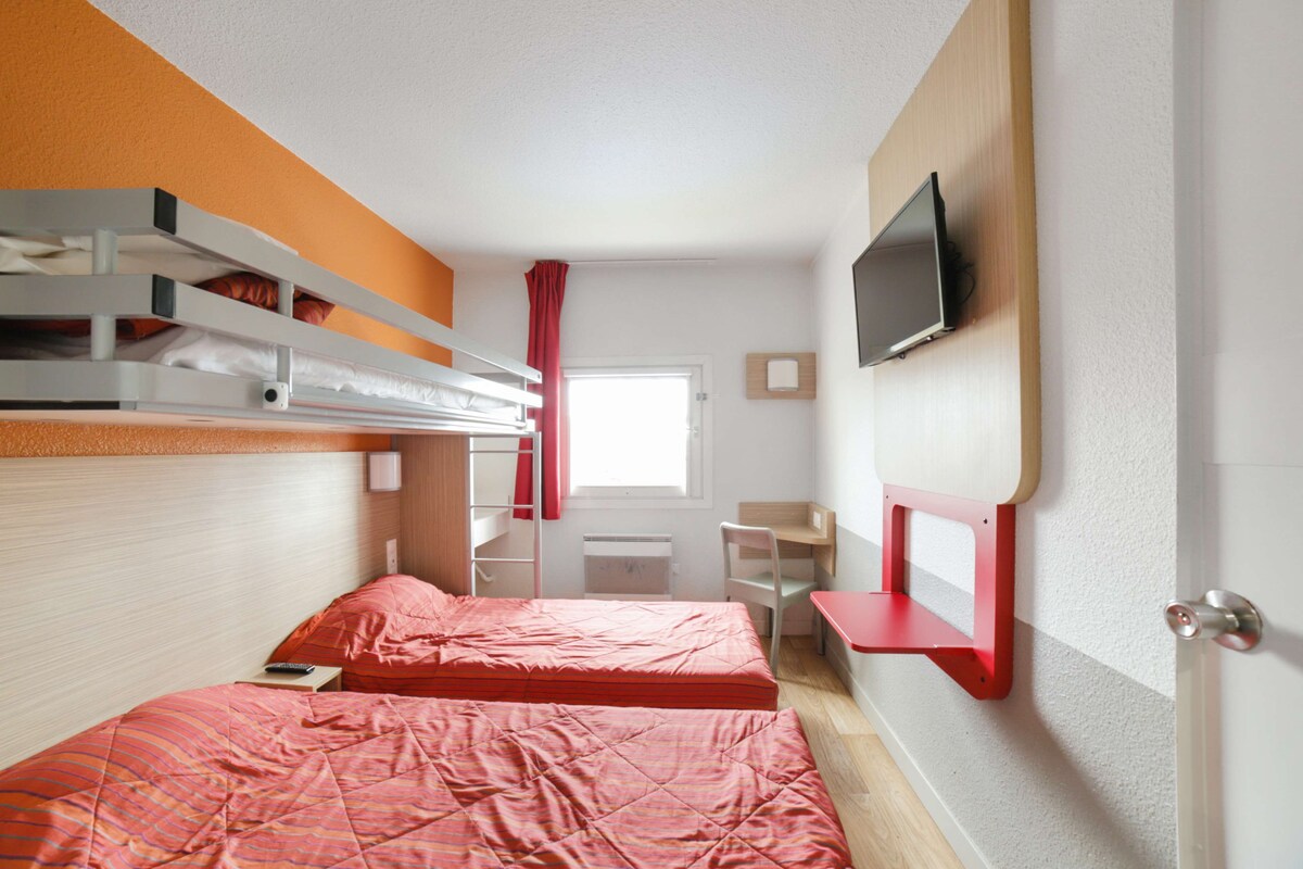 Une chambre de dortoir avec des lits superposés, un bureau, une chaise, un téléviseur fixé au mur et une fenêtre avec des rideaux.