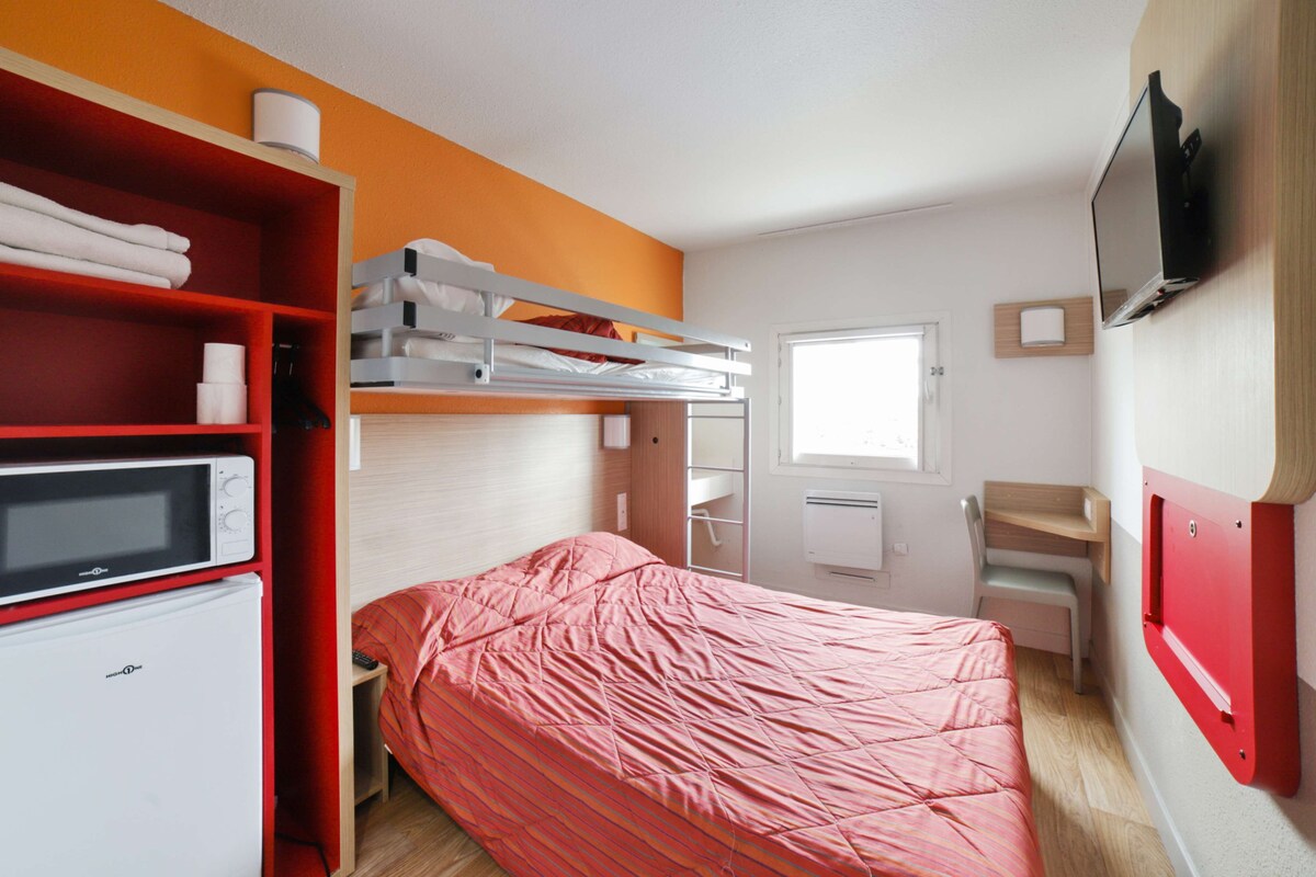 Een compacte hotelkamer met een stapelbed, een magnetron, een kleine koelkast, een aan de muur bevestigde televisie en een raam dat natuurlijk licht binnenlaat.
