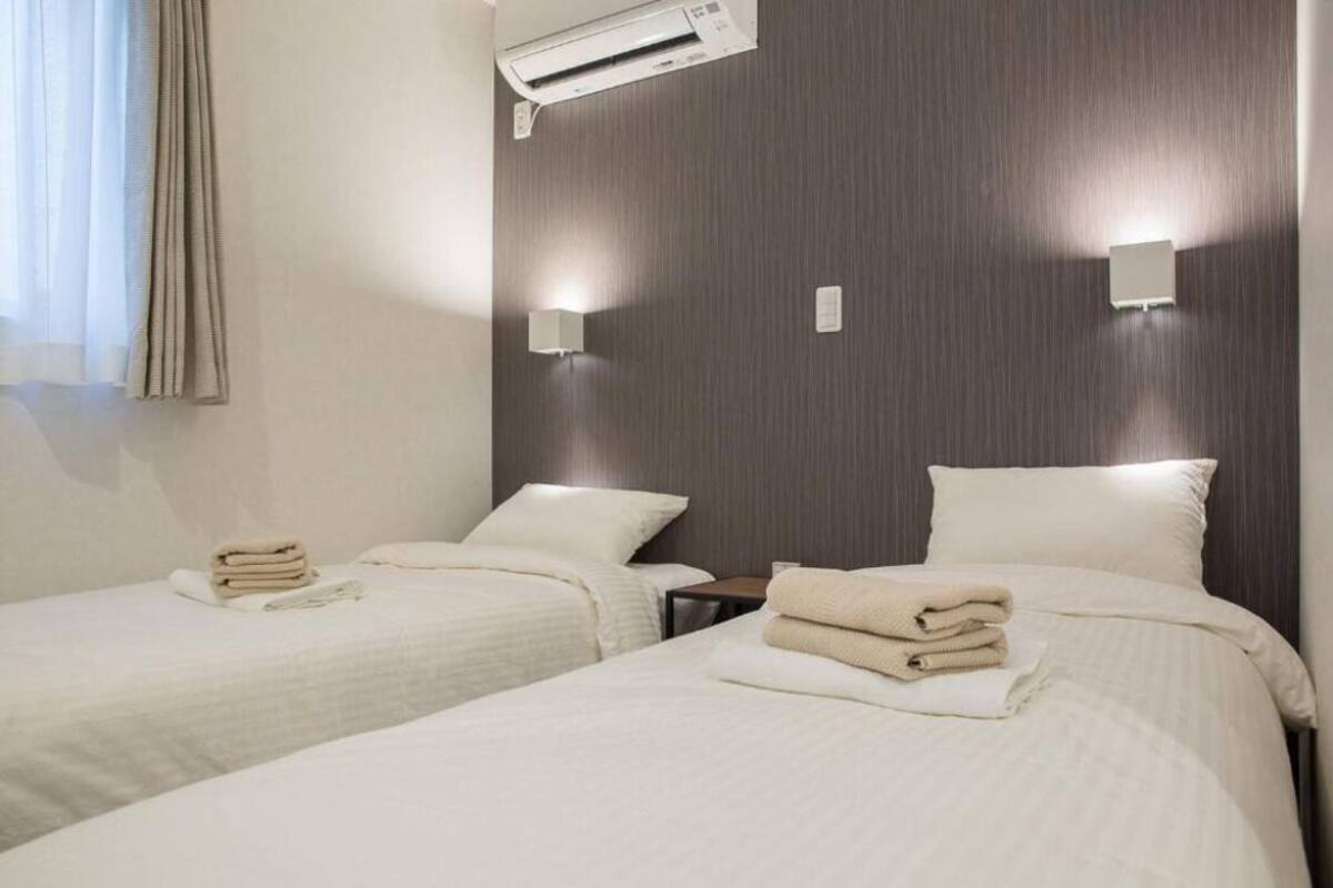 A hotel room with two beds, each with a neatly folded towel, a bedside table, and a wall-mounted air conditioning unit.