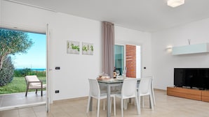 A modern dining area with a glass table, white chairs, a view of the outdoors, and a TV on a wooden cabinet. - Holiday Apartment "Villa Marconi 5" with Sea View, Covered Terrace & Large Garden (Golfo Aranci)