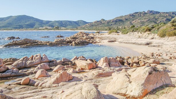 A rocky beach with clear blue water and distant mountains.