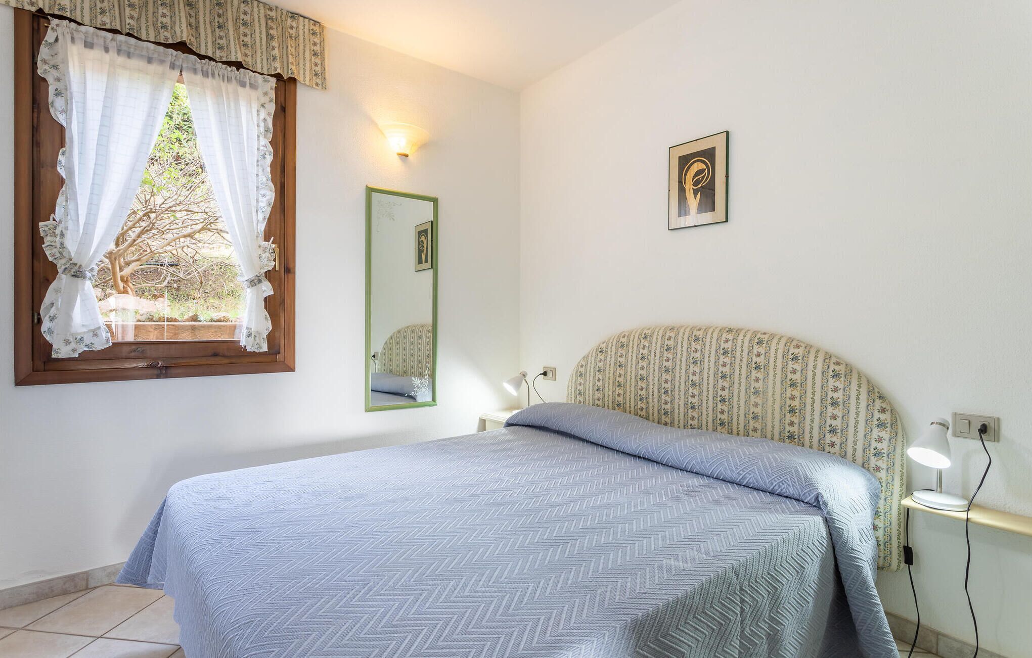 Ein Schlafzimmer mit Bett, einem Fenster mit Vorhängen, einer Nachttischlampe, einem Spiegel und einem an der Wand hängenden Bild.