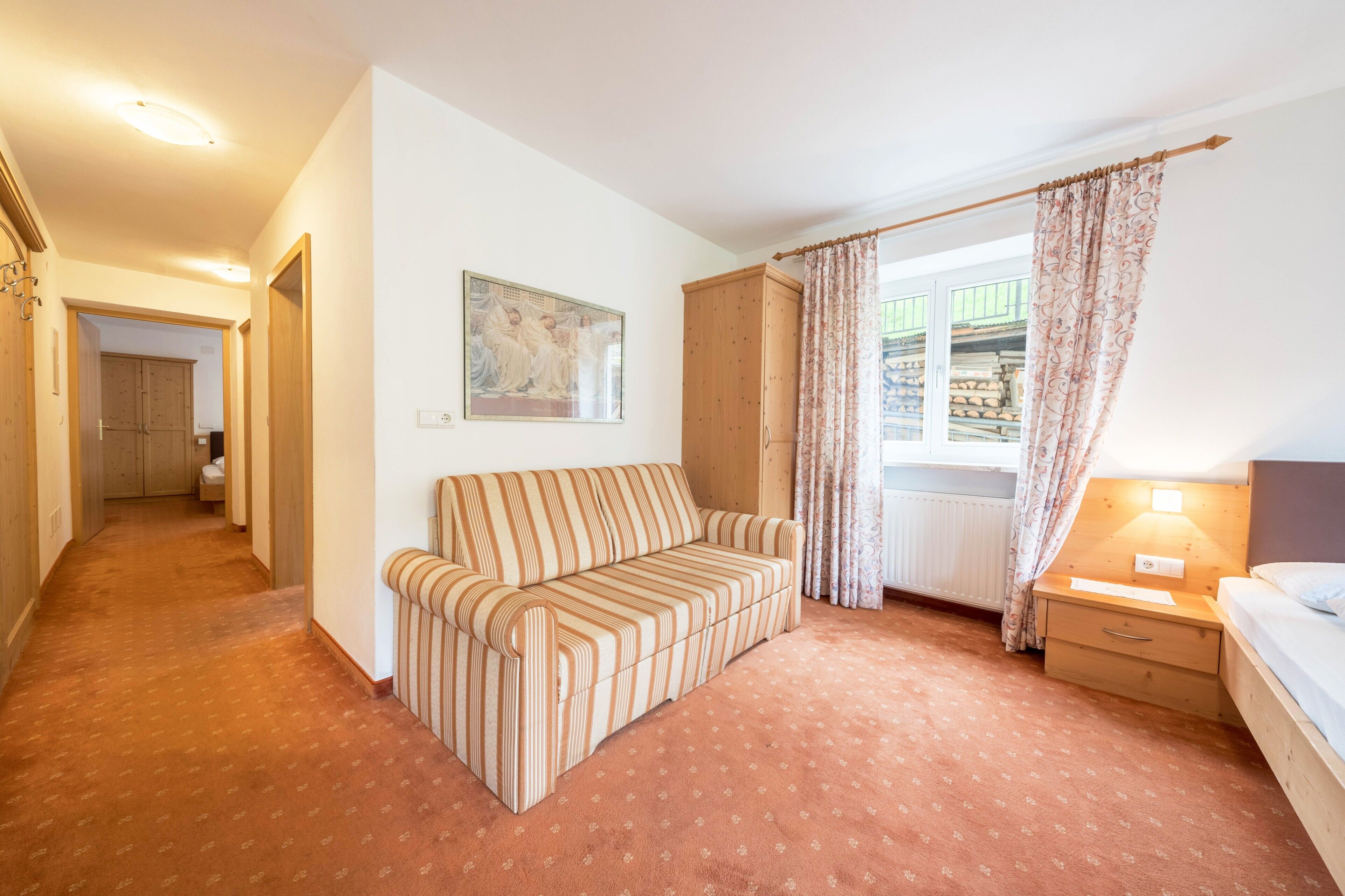 A hotel room with a striped sofa, a wooden wardrobe, a nightstand with a lamp, a bed, a window with curtains, and a hallway leading to another room.