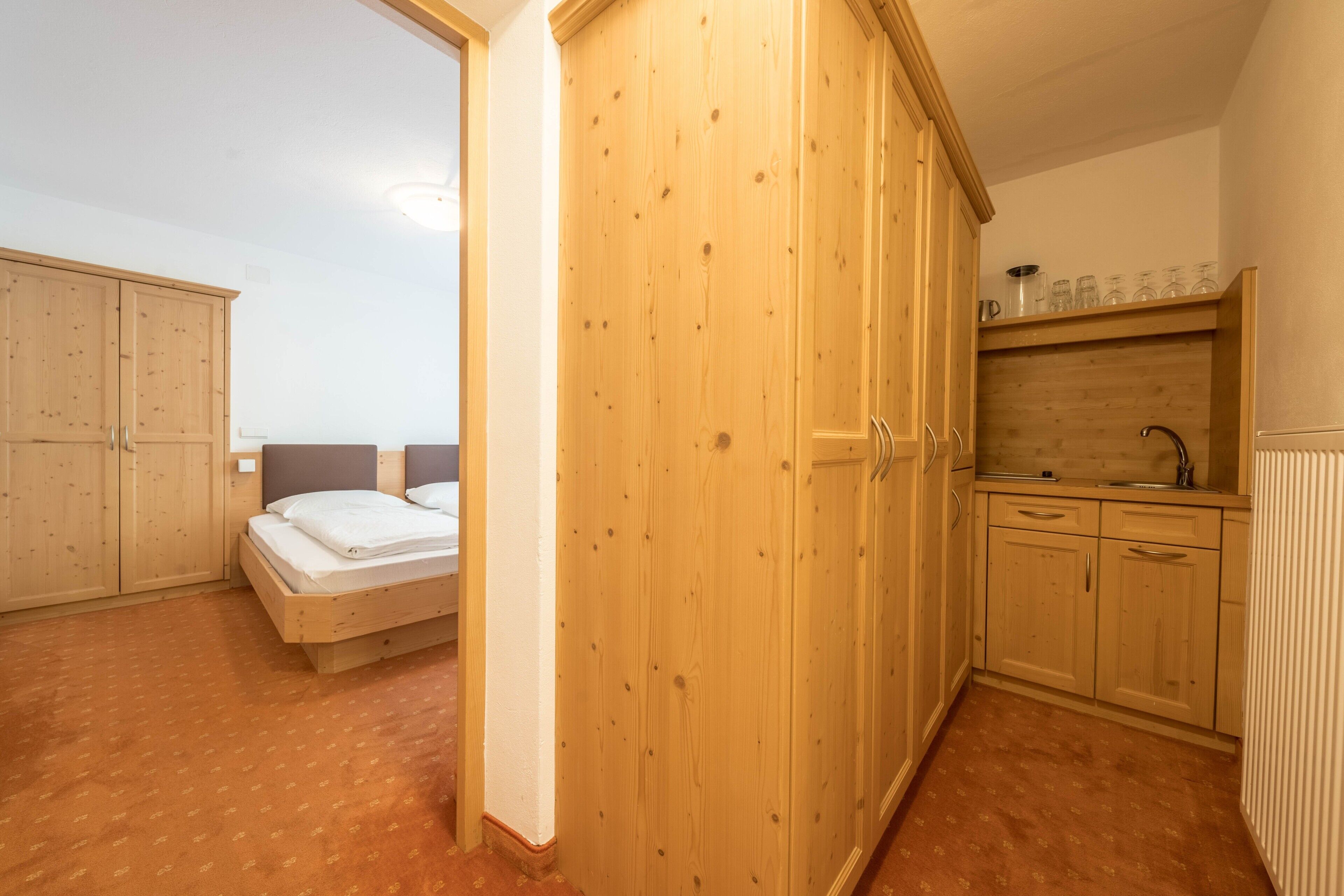 A bedroom with a bed, wooden wardrobe, and a kitchenette with a sink and cabinets.
